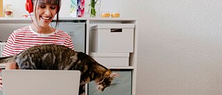 Eine junge Frau mit Laptop und rotem Kopfhörer lächelt, während eine Katze über die Tastatur geht