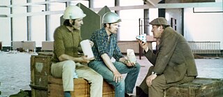A group of contruction workers wearing helmets and another man sitting an talking