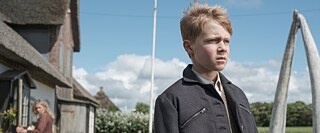 A close-up of a boy with light hair and a dark jacket looking toward the right edge of the picture, with a meadow, trees, and blue sky in the background.