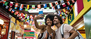 Ein junges Paar besucht einen überdachten Markt und macht ein Selfie. Über ihnen hängen viele internationale Flaggen.