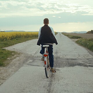 Von hinten ist eine Person zu sehen, die mit dem Fahrrad durch blühende Felder fährt.