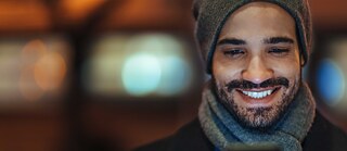 A man wearing a hat and scarf looks at his smartphone and smiles.