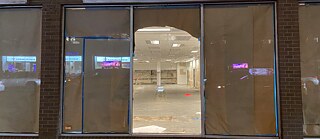 Eine teils mit Papier abgeklebte Fensterfront, durch die ein leerer Innenraum mit hellen Deckenlichtern sichtbar ist.