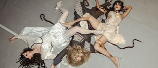 Kolm pikkade juustega naist lamavad maas. Nende käed ja jalad on üksteisega põimunud.