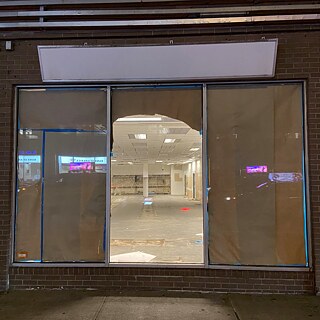 Eine teils mit Papier abgeklebte Fensterfront, durch die ein leerer Innenraum mit hellen Deckenlichtern sichtbar ist.