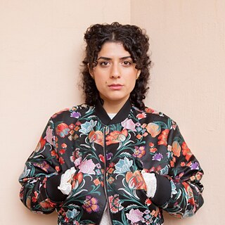 Eine Frau mit geblümter Jacke und weißem Hemd steht vor einer rosa Wand.