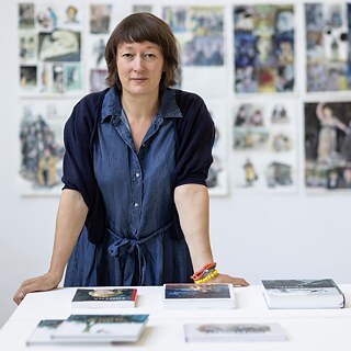 Barbara Yelin steht hinter einem Schreibtisch; hinter ihr eine Wand mit verschiedenen Illustrationen der Künstlerin.