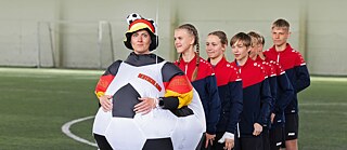 Mokytoja su futbolo kostiumu stovi prieš eilę jaunuolių sportine apranga futbolo aikštėje.