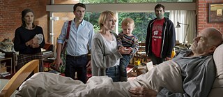 In einem Zimmer betrachten eine junge Frau mit einem Kind auf dem Arm, eine ältere Frau und zwei Männer einen älteren Mann, der in einem Krankenhausbett liegt.