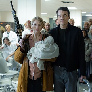 Eine junge Frau, die in einer Hand ein Gewehr hält und auf dem anderen Arm ein Baby trägt, steht neben einem jungen Mann. Im Hintergrund sehen wir einige Menschen in Krankenhauskleidung.