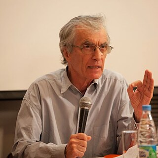 Das Bild zeigt eine Person, die an einem Tisch sitzt und offenbar an einer Veranstaltung oder Diskussion teilnimmt. Die Person trägt ein helles, langärmliges Hemd und hält in der linken Hand ein Mikrofon, als würde gerade gesprochen oder eine Antwort gegeben. Die rechte Hand ist erhoben, mit Daumen und Zeigefinger zu einem Kreis geformt, was oft eine Geste zur Betonung oder Erklärung ist. Auf dem Tisch vor der Person stehen mehrere Wasserflaschen mit blauen Verschlusskappen, die in einer Reihe angeordnet sind. Der Hintergrund ist schlicht und hell, vermutlich eine Projektionsfläche oder Wand.