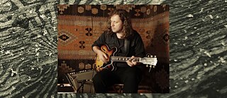 Auf dem Foto ist der Gitarrist Raphael Rogiński zu sehen, der im Sitzen eine halbakustische E-Gitarre spielt und sich ganz auf sein Instrument konzentriert. Der warme, textile Hintergrund und das gedämpfte Licht erzeugen eine intime Atmosphäre, die an eine Probe oder eine Aufnahme im kleinen Rahmen erinnert. 