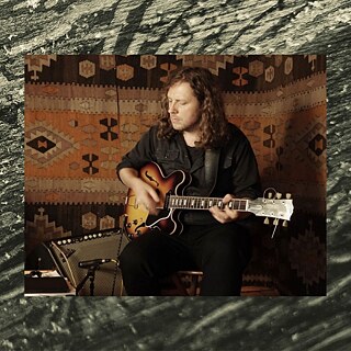 Auf dem Foto ist der Gitarrist Raphael Rogiński zu sehen, der im Sitzen eine halbakustische E-Gitarre spielt und sich ganz auf sein Instrument konzentriert. Der warme, textile Hintergrund und das gedämpfte Licht erzeugen eine intime Atmosphäre, die an eine Probe oder eine Aufnahme im kleinen Rahmen erinnert. 