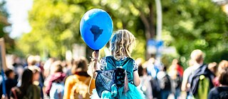 A young girl with a balloon signifying the earth sits on the shoulder of an adult. A crowd marching for climate justice.