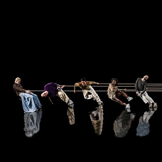 5 Tänzer*innen in einer Reihe auf einer komplett schwarzen Bühne, zwischen ihnen sind Seile gespannt und ihre Silhouetten spiegeln sich im Boden.