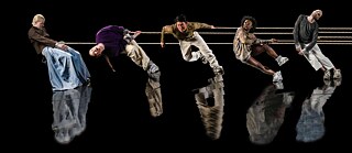 5 Tänzer*innen in einer Reihe auf einer komplett schwarzen Bühne, zwischen ihnen sind Seile gespannt und ihre Silhouetten spiegeln sich im Boden.