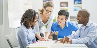 Vier Personen, zwei Frauen und zwei Männer sitzen um einen Schreibrich herum. Eine Frau, die steht, hält einen Stift in der rechten Hand und notiert etwas auf einem Blatt, auf das die anderen drei Personen schauen. Die Farben des Fotos sind sehr hell. 