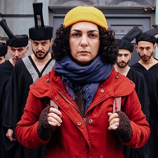 Szene aus dem Film SHAHID: Eine Frau in gelber Mütze und roter Jacke steht vor einer Gruppe schwarz gekleideter Männer.