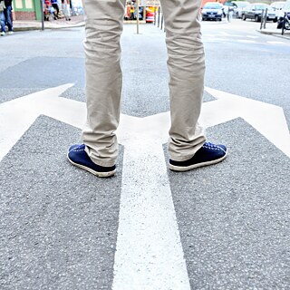 Person steht auf einer Straßenmarkierung in Form eines großen Pfeils, der nach links und rechts zeigt. Die Person trägt beige Hosen und dunkelblaue Schuhe. Im Hintergrund sind Autos und Gebäude auf einer Stadtstraße zu sehen.