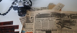 Eine Vintage-Kamera, Filmnegative und verstreute alte Zeitungsausschnitte liegen auf einem Tisch als Teil eines GPS‑2026‑Archivprojekts.