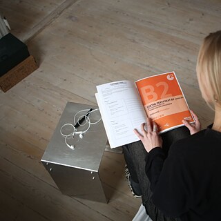 Eine Deutschlehrerin sitzt vor einem Computer und hält eine Goethe-Prüfung B2 in den Händen.