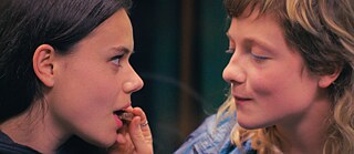 Δύο γυναίκες κάθονται κοντά η μία στην άλλη, η μία δίνει στην άλλη κάτι να φάει. Η σκηνή φαίνεται οικεία και δείχνει μια στενή επαφή.