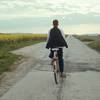 Ένας αγόρι ποδηλατεί σε ένα ευθύ αγροτικό δρόμο, πλαισιωμένο από κίτρινα χωράφια και χέρσα γη κάτω από έναν συννεφιασμένο ουρανό.