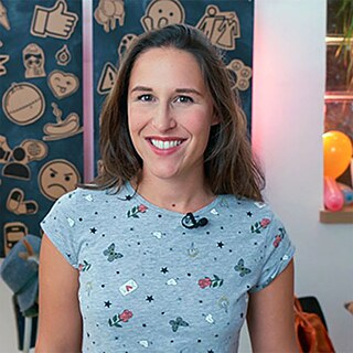 Eine junge Frau steht lächelnd im Zentrum, im Hintergrund eine dekorierte Wand und ein Fenster