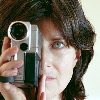 Eine Person hält eine kleine, silberfarbene Videokamera nah vor ihrem Gesicht. Die Kamera ist im Vordergrund deutlich sichtbar, mit einem großen Objektiv und einem roten Aufnahme‑Lämpchen. Die Person umfasst das Gerät mit einer Hand; die Finger sind am unteren Rand des Kameragehäuses zu sehen