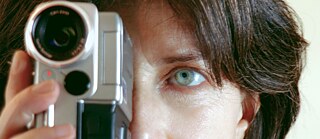 Eine Person hält eine kleine, silberfarbene Videokamera nah vor ihrem Gesicht. Die Kamera ist im Vordergrund deutlich sichtbar, mit einem großen Objektiv und einem roten Aufnahme‑Lämpchen. Die Person umfasst das Gerät mit einer Hand; die Finger sind am unteren Rand des Kameragehäuses zu sehen