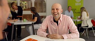 Ein Mann sitzt an einem Tisch und führt lächelnd ein Gespräch.