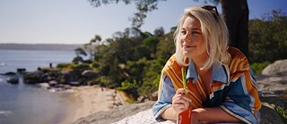 Eine Frau liegt vor einem Notizbuch auf einem Stein und genießt den Meerblick während sie einen Stift hält.