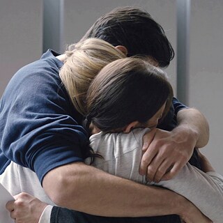 Τρεις άνθρωποι αγκαλιάζονται σφιχτά σε ένα φωτεινό δωμάτιο, κοντινό πλάνο της πλάτης και των χεριών τους.