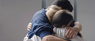 Τρεις άνθρωποι αγκαλιάζονται σφιχτά σε ένα φωτεινό δωμάτιο, κοντινό πλάνο της πλάτης και των χεριών τους.