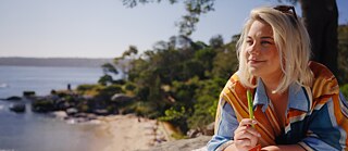 Eine Frau liegt vor einem Notizbuch auf einem Stein und genießt den Meerblick während sie einen Stift hält.