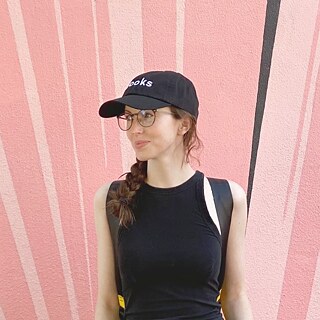 Die Autorin Lena Anlauf steht in einem schwarzen, ärmellosen Top und mit Schirmmütze vor einer rosafarbenen Wand.