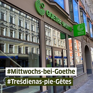Fassade des Goethe-Instituts in Marijas Straße 13 k1 mit großen Schaufenstern, in denen sich gegenüberliegende Gebäude spiegeln. Über dem Eingang hängt das grüne Logo des Goethe-Instituts. Im Vordergrund stehen die Hashtags „#Mittwochs-bei-Goethe“ und „#Trešdienās-pie-Gētes“ in weißen und grünen Textbalken.