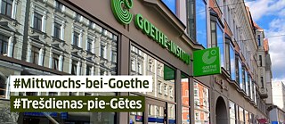 Fassade des Goethe-Instituts in Marijas Straße 13 k1 mit großen Schaufenstern, in denen sich gegenüberliegende Gebäude spiegeln. Über dem Eingang hängt das grüne Logo des Goethe-Instituts. Im Vordergrund stehen die Hashtags „#Mittwochs-bei-Goethe“ und „#Trešdienās-pie-Gētes“ in weißen und grünen Textbalken.
