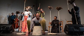 Performance scene from Selemo by Sbusiso Shozi and Nhlanhla Mahlangu at the Centre for the Less Good Idea, showing performers on stage holding feathered sticks while one person kneels in front with woven baskets and bundles of grass.