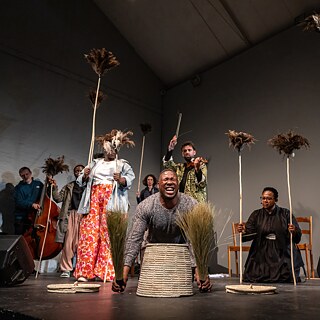 Performance scene from Selemo by Sbusiso Shozi and Nhlanhla Mahlangu at the Centre for the Less Good Idea, showing performers on stage holding feathered sticks while one person kneels in front with woven baskets and bundles of grass.