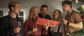 Five people with champagne glasses stand solemnly together and sing from a book. 