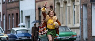 Μια νεαρή γυναίκα περπατά σε έναν βρεγμένο δρόμο σε μια πόλη με ιστορικά κτίρια. Φορώντας ένα πολύχρωμα ρετρό ρούχα και κρατά μια τσάντα με μοτίβα. Στο βάθος φαίνονται κλασικά αυτοκίνητα και περαστικοί με ομπρέλες.