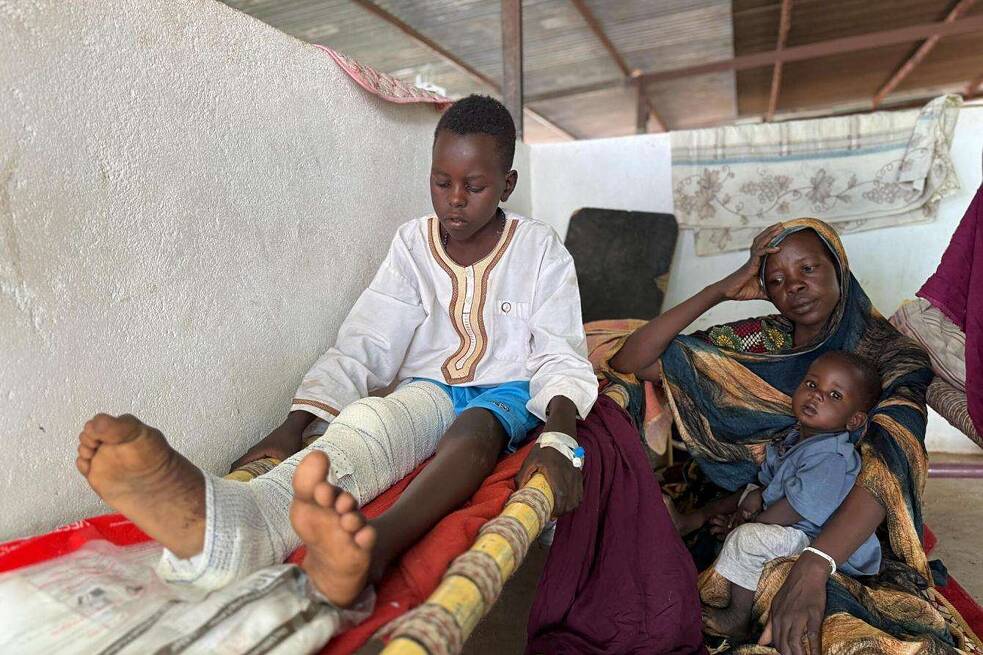 Ein Junge sitzt auf einer Pritsche in einem Zelt. Sein rechtes Bein ist verletzt. Neben ihn sitzt eine Frau mit Hijab. Sie hat ein Kind auf dem Schoß und sieht müde aus.