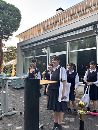 この写真は、東京にあるドイツ大使館の庭園で、演台の前に立つ女子学生を写しています。