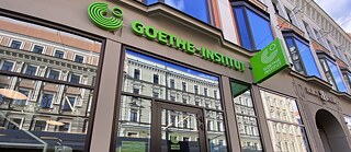 Moderne Glasfassade des Goethe-Instituts Riga mit grünem Logo und Schriftzug über dem Eingang; in den großen Fenstern spiegelt sich die gegenüberliegende historische Häuserzeile.