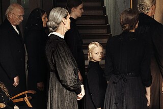 A little girl with blonde pigtails looks sadly into the camera. She is surrounded by family members dressed in mourning clothes. 