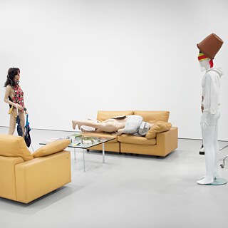 En installation viser flere dukkelignende figurer i et hvidt gallerirum, arrangeret omkring to gule sofaer og et glasbord med spredte genstande. En figur ligger udstrakt på sofaen, mens to andre står oprejst ved siden af, heriblandt en med en blomsterpotte over hovedet.