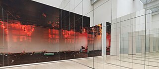 En installation viser et stort vægprint af et forladt, rødt oplyst rum med spredte genstande, kombineret med omgivende, gulvhøje spejle. Spejlene forstørrer rummet og skaber en labyrintisk, forvrænget perspektiv.