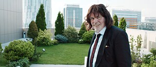 Ein Mann im Rentenalter mit Perücke und falschen Zähnen steht im Anzug gekleidet auf einer Dachterasse.