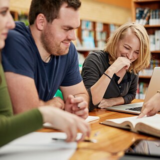 Drei Personen arbeiten gemeinsam an etwas in einer Bibliothek, sie lachen.